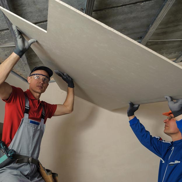 Two workers positioning a sheet of drywall against a ceiling